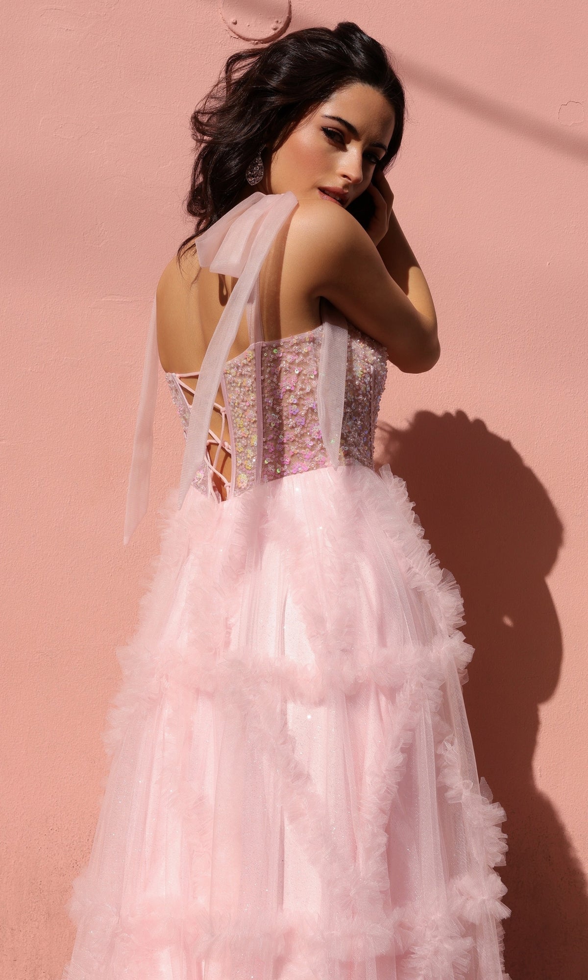 Woman in the Shoulder-Bows Long Ruffled Prom Gown T1340—pink, sleeveless tulle with beaded corset bodice and lace-up back—stands against a pink wall, glancing over her shoulder.
